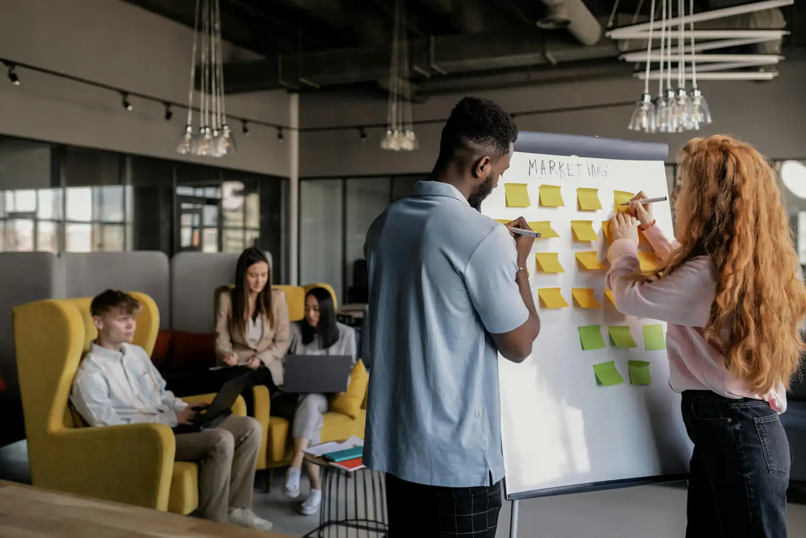 Marketing team writing campaign strategy on whiteboard with sticky notes in modern office
